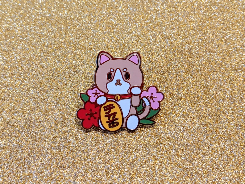 May include: A brown and white cat with a red collar and a gold coin with Japanese characters that reads "lucky cat" in English. The cat is holding a pink flower in its paw and is surrounded by pink flowers and green leaves.