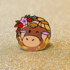 May include: A gold-tone enamel pin featuring a cartoon ox with a pink nose and brown fur. The ox is wearing a wreath of red and pink flowers and has the year 2021 printed below it.