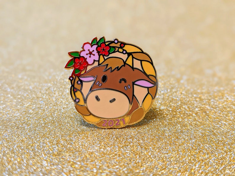 May include: A gold-tone enamel pin featuring a cartoon ox with a pink nose and brown fur. The ox is wearing a wreath of red and pink flowers and has the year 2021 printed below it.