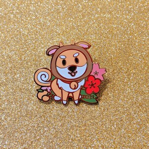 May include: A gold enamel pin featuring a cartoon dog wearing a red and white scarf and holding a pink flower. The dog is surrounded by pink flowers and green leaves. The pin says "2021" on the bottom.