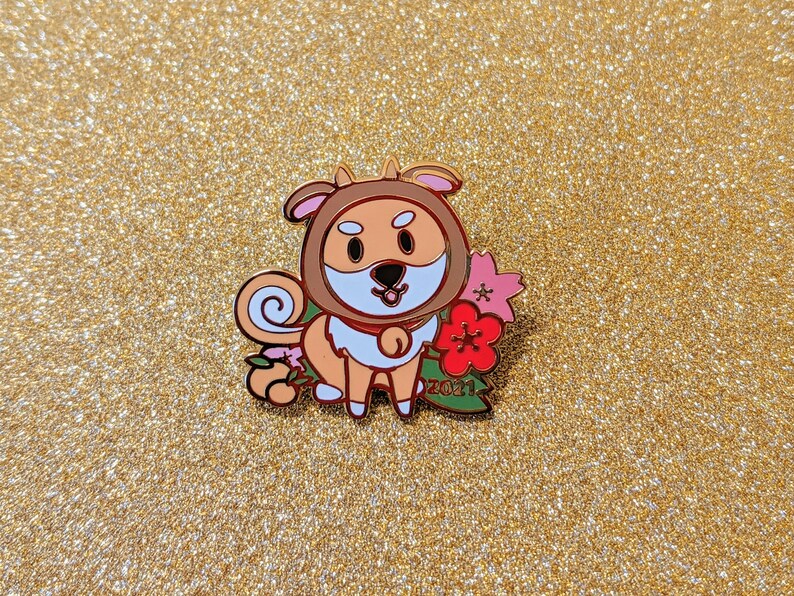 May include: A gold enamel pin featuring a cartoon dog wearing a red and white scarf and holding a pink flower. The dog is surrounded by pink flowers and green leaves. The pin says "2021" on the bottom.