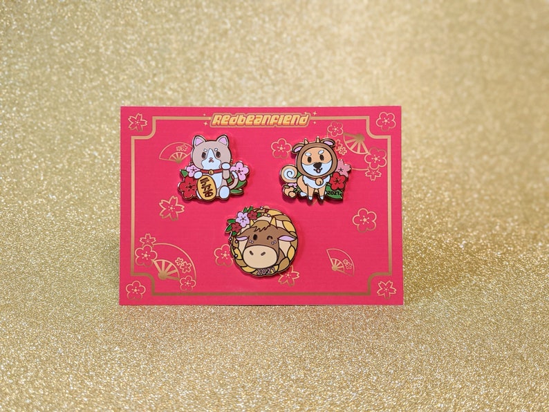 May include: Three enamel pins featuring cartoon animals on a red background. The pins are part of a set and feature a cat, a dog, and a cow. The pins are surrounded by pink flowers and gold accents.