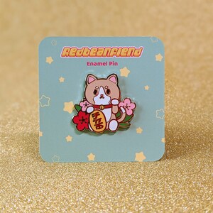 May include: A gold enamel pin featuring a cartoon cat holding a red and gold lucky charm with the Chinese characters for "good luck" written on it. The cat is surrounded by pink and red flowers. The pin is on a blue background with white stars.