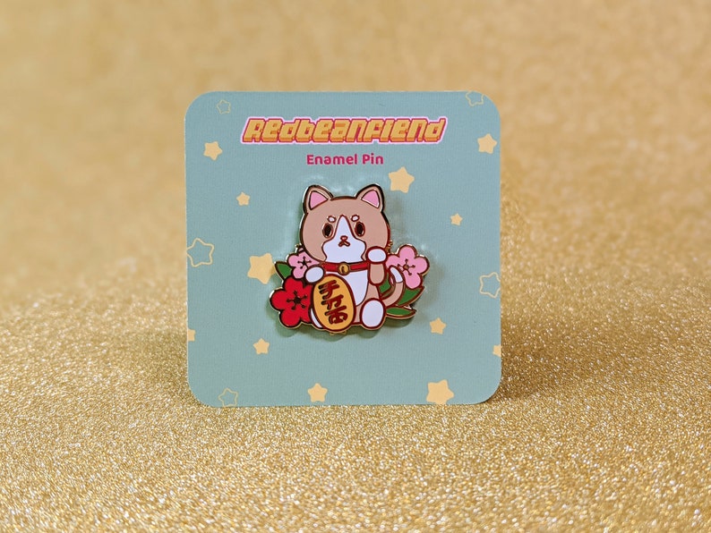 May include: A gold enamel pin featuring a cartoon cat holding a red and gold lucky charm with the Chinese characters for "good luck" written on it. The cat is surrounded by pink and red flowers. The pin is on a blue background with white stars.