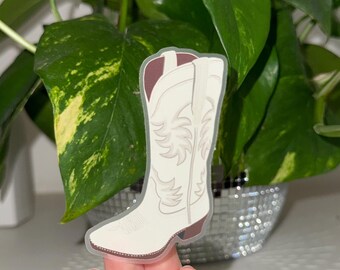 Op de afbeelding: Een witte cowboylaarzensticker met bruine accenten. De sticker toont een gedetailleerd ontwerp van een klassieke westernlaars, met decoratieve stiksels en een puntige neus. De laars staat tegen een vage achtergrond van groene bladeren.