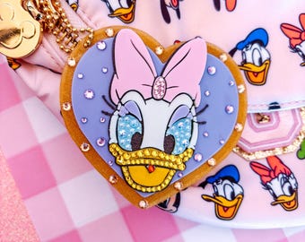 May include: A pink fanny pack featuring a repeating pattern of Disney characters. A heart-shaped charm with Daisy Duck's face is attached. The charm is decorated with rhinestones and a pink bow. A gold-colored zipper and hardware are visible.