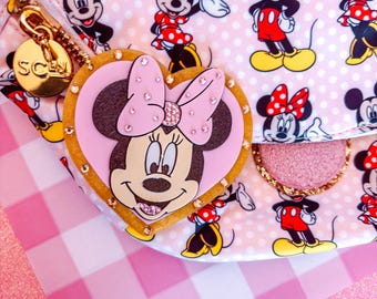 May include: Pink fanny pack featuring a repeating pattern of Mickey Mouse and Minnie Mouse. The bag has a gold zipper and a heart-shaped Minnie Mouse charm with rhinestones. The background is a pink and white gingham pattern.