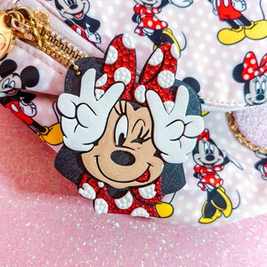May include: A Minnie Mouse-themed accessory with a black silhouette, red polka dot bow, and white hands making a peace sign. The accessory is set against a pink background with a pattern of Minnie and Mickey Mouse characters.