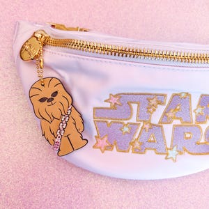 May include: A white fanny pack with a gold zipper and a gold chain attached to a brown Chewbacca charm. The fanny pack has the words "STAR WARS" embroidered in purple with gold glitter stars.