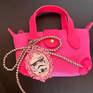 May include: A bright pink handbag with a top handle and a decorative charm. The charm features a framed image of a Stormtrooper helmet. The bag has a flap closure with a gold-colored button.