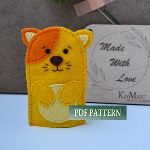 May include: A felt finger puppet of a yellow cat with orange ears and a light yellow belly. The cat has a brown nose and black eyes. The puppet is sitting on a white surface with a wooden sign that says "Made with Love" and "KsyMart" in the background. The sign also has a floral design. The text "PDF PATTERN" is printed on the surface below the puppet.