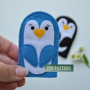 Könnte beinhalten: Blau-weiße Filz-Fingerpuppe in Form eines Pinguins mit gelbem Schnabel und schwarzen Augen. Die Puppe wird von einer Hand gehalten. Der Text "PDF PATTERN" ist im Bild sichtbar.