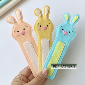 May include: Three felt bookmarks shaped like rabbits in peach, yellow, and light blue. Each rabbit has stitched facial features, pink noses, and contrasting inner ear and belly patches. The text PDF PATTERN is visible on the blue bookmark.