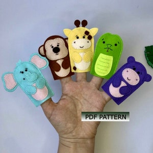 May include: Five felt finger puppets in the shape of animals: an elephant, monkey, giraffe, crocodile, and hippopotamus. The puppets are all smiling and have stitched details. The puppets are on a hand, with the text "PDF PATTERN" below.