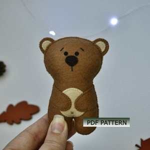 May include: A brown felt bear with black eyes and a black nose. The bear is standing on a hand. The text "PDF PATTERN" is visible in the lower right corner of the image.