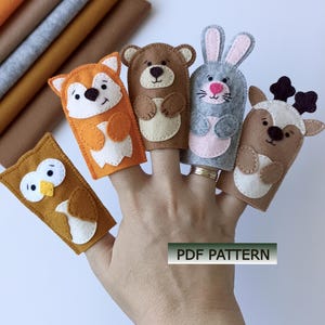 May include: A set of five felt finger puppets featuring an owl, a fox, a bear, a rabbit, and a deer, each with distinct facial features and colors. The puppets are displayed on fingers, and a rectangular label with the text PDF PATTERN is visible.