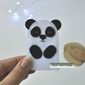 May include: A white felt panda finger puppet with black ears, eyes, and belly. The puppet has a smiling face and is held by a hand. The text "PDF PATTERN" is visible on the puppet.