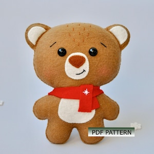 May include: A handmade brown teddy bear with a red scarf featuring a white star. The bear has black button eyes, a brown nose, and white accents on its ears and belly. The text "PDF PATTERN" is visible.