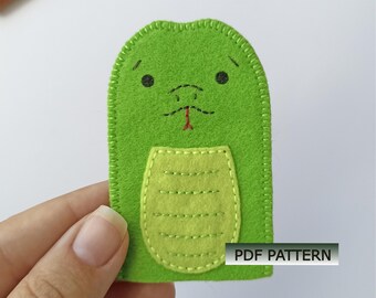 May include: A green felt finger puppet shaped like a snake with a light green belly patch, embroidered black eyes, a small mouth, and a red forked tongue. The edges are finished with a blanket stitch. The image includes the text PDF PATTERN.