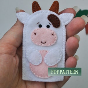 Puede incluir: Marionetas de dedo de fieltro blanco de una vaca con orejas marrones, una mancha marrón, nariz rosa y ojos negros. La marioneta tiene una barriga rosa y pezuñas blancas. El texto "PDF PATTERN" es visible en la imagen.