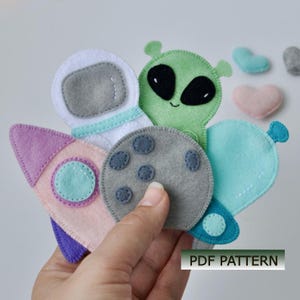 May include: A handmade felt space-themed toy set featuring a rocket, astronaut, alien, moon, and heart shapes in pastel colors. The rocket is pink and purple, the alien is green, and the moon is gray. The text "PDF PATTERN" is visible.