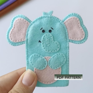 May include: A light blue felt finger puppet shaped like an elephant with light pink inner ears and a light pink belly. The elephant features stitched facial details, including eyes and a trunk, and includes the text PDF PATTERN on the lower right.