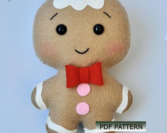 May include: A felt gingerbread man doll with a tan body, white frosting, and a red bow tie. The doll has black button eyes, rosy cheeks, and a smiling mouth. White accents are on the arms and legs. The text "PDF PATTERN" is visible.