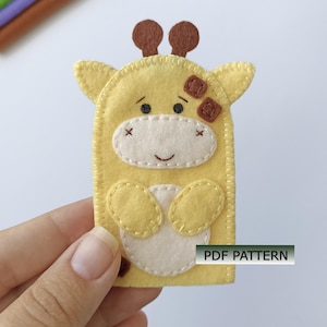 Könnte beinhalten: Eine gelbe Fingerpuppe aus Filz in Form einer Giraffe mit braunen Hornzapfen, einer cremefarbenen Schnauze und einem cremefarbenen Bauchfleck. Die Puppe hat gestickte Details, kleine dunkle Augen und ein Lächeln. Ein Etikett zeigt PDF PATTERN.