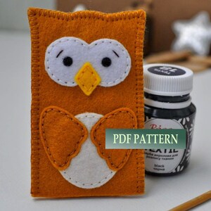 May include: A felt owl craft project with a PDF pattern. The owl is orange with white eyes and a yellow beak. The owl is sitting on a white surface next to a bottle of black fabric paint.