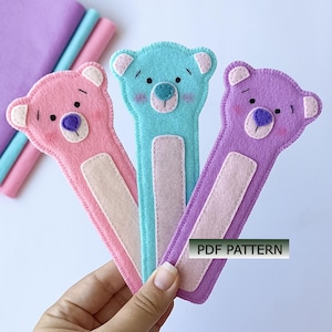 May include: Three felt bear bookmarks in pink, light blue, and purple. Each bookmark features a bear face with stitched details, a light pink belly patch, and a rectangular label with the text PDF PATTERN printed in black on a white background.