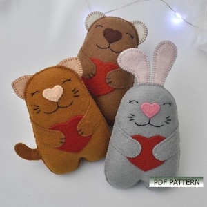 Può includere: Tre peluche di animali in feltro che tengono cuori rossi. Un gatto marrone, un orso marrone e un coniglio grigio.  MODELLO PDF.