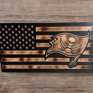 May include: Wooden American flag with the Tampa Bay Buccaneers logo. The flag features a distressed design with the team's logo replacing the traditional stars and stripes. The flag is made of wood and has a dark finish.