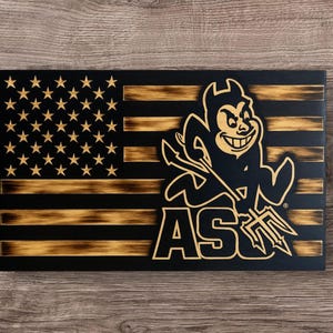 Op de afbeelding: Een houten Amerikaanse vlag met een zwarte en gouden afwerking. De vlag toont de mascotte van de Arizona State University en de letters "AS" in goud. De vlag is rechthoekig en heeft een verweerde uitstraling.