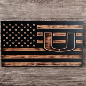 Könnte beinhalten: Eine amerikanische Flagge aus Holz mit einem Used-Look. Die Flagge zeigt das Sternen- und Streifenmuster mit einem großen Buchstaben "U" in der Mitte. Die Sterne befinden sich in der oberen linken Ecke, und die Streifen sind horizontal.