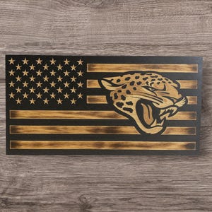 May include: A wooden American flag with a black background and gold stripes. The stars are gold, and a gold jaguar head is on the right side. The flag is rectangular and has a distressed finish.