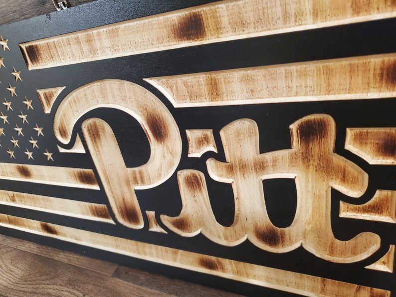 May include: A wooden sign with a black background and a distressed wood finish. The sign features a stylized American flag with the word "Pitt" in a large, white font.