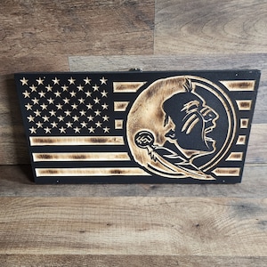 May include: A black wooden sign featuring the American flag on the left and a Seminole head logo on the right. The flag has carved stars and stripes. The Seminole logo is in a circle with a detailed profile.