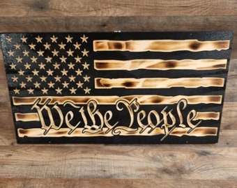 Puede incluir: Una bandera americana de madera con fondo negro y las palabras "We the People" quemadas en la madera. La bandera está desgastada y tiene un aspecto rústico.