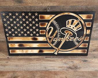 May include: A wooden sign with a black background and a burnt wood effect. The sign features a stylized image of the New York Yankees logo with a baseball and bats. The logo is surrounded by a circle with the word "Yankees" written inside. The sign also features a small American flag in the top left corner.