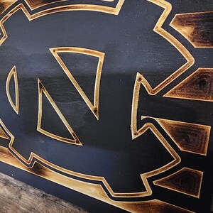 Pode incluir: Uma placa de madeira com um design de bandeira americana queimada e um contorno dourado do logotipo dos Tar Heels da Universidade da Carolina do Norte.