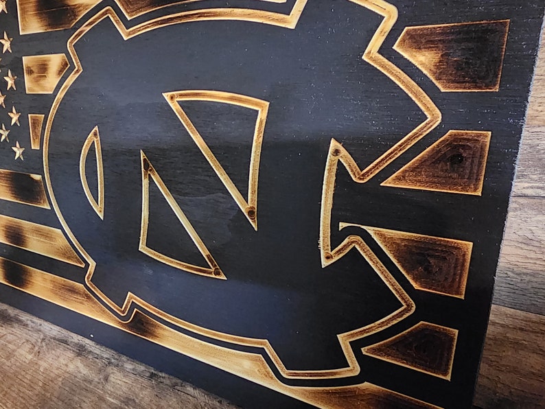 Pode incluir: Uma placa de madeira com um design de bandeira americana queimada e um contorno dourado do logotipo dos Tar Heels da Universidade da Carolina do Norte.