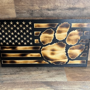 May include: A wooden American flag with a burnt wood design. The flag has a paw print in the centre, also with a burnt wood design.