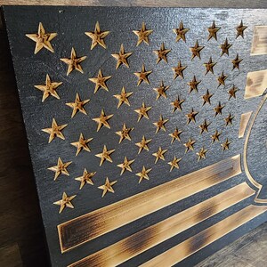 Pode incluir: Uma bandeira americana de madeira com um visual desgastado e queimado. As estrelas s&atilde;o douradas e as listras s&atilde;o pretas e marrons. A bandeira tem uma grande letra "C" estilizada preta no canto inferior direito.