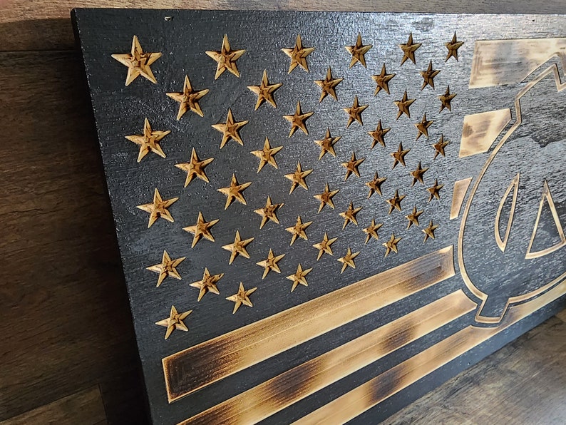 Pode incluir: Uma bandeira americana de madeira com um visual desgastado e queimado. As estrelas s&atilde;o douradas e as listras s&atilde;o pretas e marrons. A bandeira tem uma grande letra "C" estilizada preta no canto inferior direito.