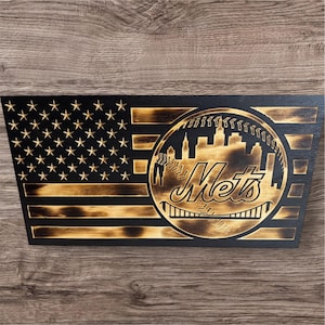 May include: Wooden American flag with a baseball design. The flag features the New York Mets logo, a cityscape, and the word "Mets" in a stylized font. The stars and stripes are burned into the wood, creating a rustic look.