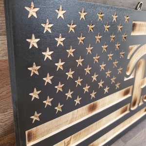 May include: A wooden American flag with a distressed look, featuring a black background and white stars. The flag is burned around the edges and has the word "Pitt" in a stylized font on the right side.
