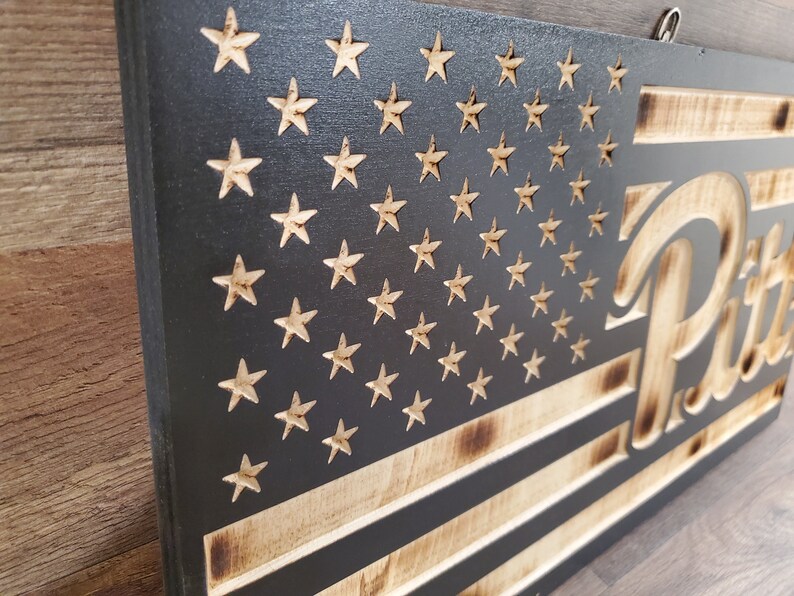 May include: A wooden American flag with a distressed look, featuring a black background and white stars. The flag is burned around the edges and has the word "Pitt" in a stylized font on the right side.
