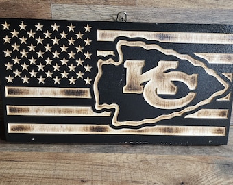 May include: A wooden sign with a black background and a distressed American flag design. The flag has a white star pattern and red and white stripes. The sign also features a carved Kansas City Chiefs logo in the centre.