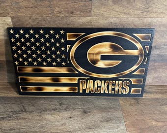May include: A wooden sign with a distressed, burnt look, featuring an American flag with the Green Bay Packers logo and the word "PACKERS" below it.