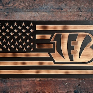May include: A rectangular wooden wall art piece featuring a burnt wood finish. The design displays a stylized American flag with stars on the left and horizontal stripes, with the Cincinnati Bengals logo integrated into the right side of the flag.